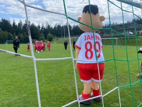 Das Maskottchen Jahni steht vor einem Tor, mit dem Rücken zur Kamera. Im Hintergrund sind eine Gruppe von Kindern in Fußballtrikots und zwei Trainer auf einem grünen Fußballfeld zu sehen.