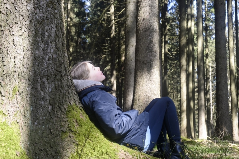 Kind lehnt sich an einen Baum und schaut in den Himmel. Im Hintergrund ist ein dicht bewachsener Wald zu erkennen.