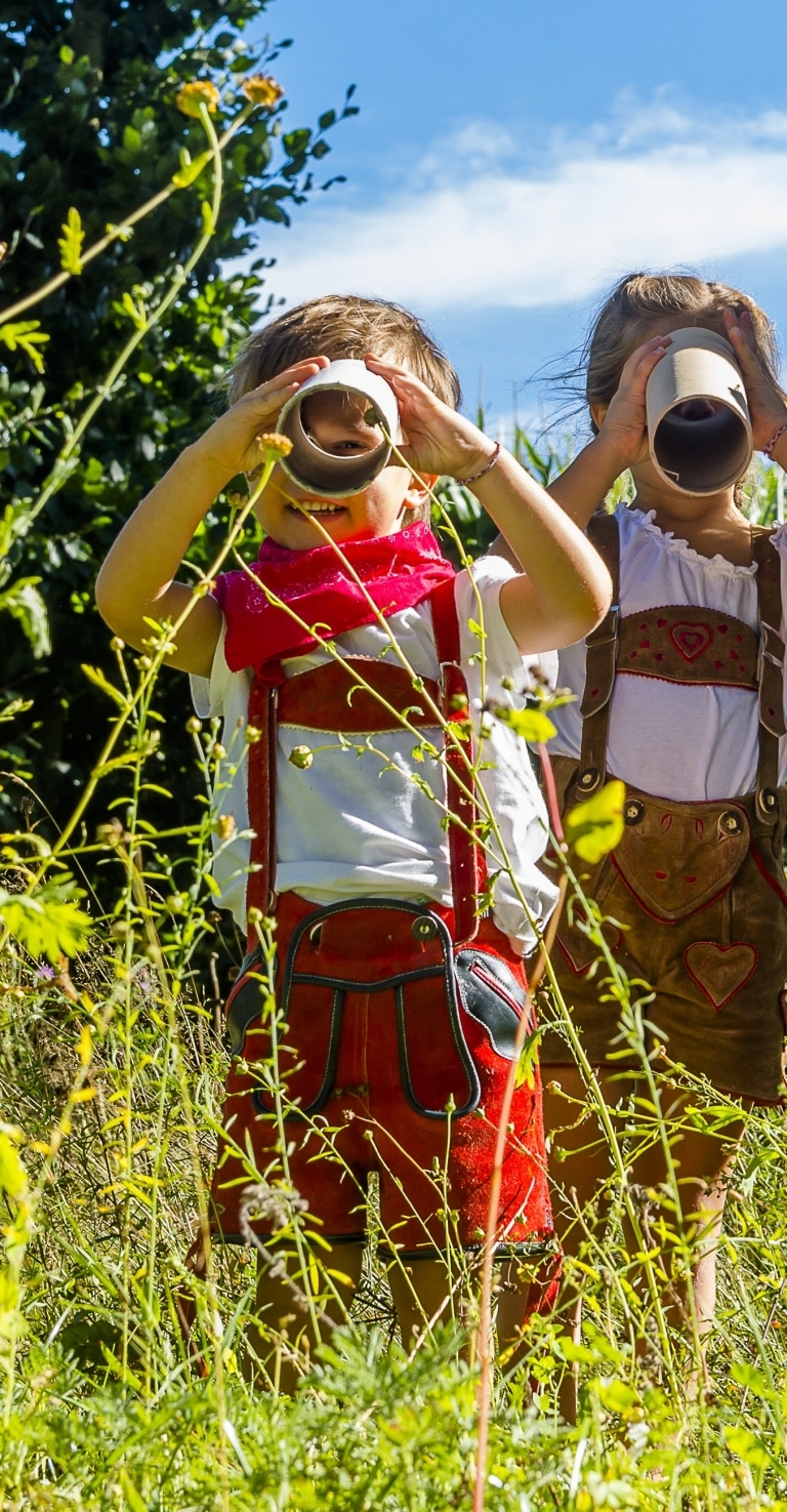 Zwei Kinder in Lederhose, spielen draußen in einem Feld, halten Pappröhren vor ihre Augen und schauen durch sie, als ob sie Ferngläser wären.