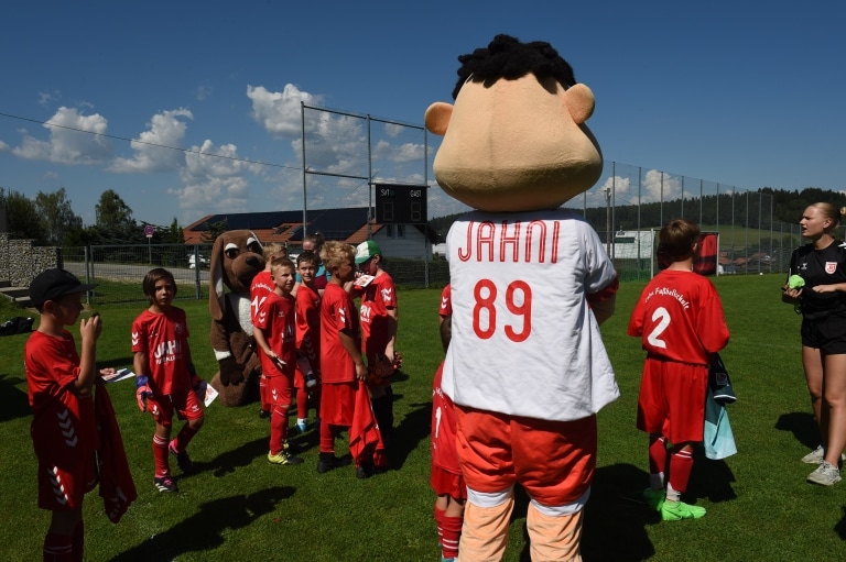 Jahni, das Maskottchen vom SSV Jahn Regensburg und Kinder in roten Fußballtrikots stehen auf dem Fußballplatz