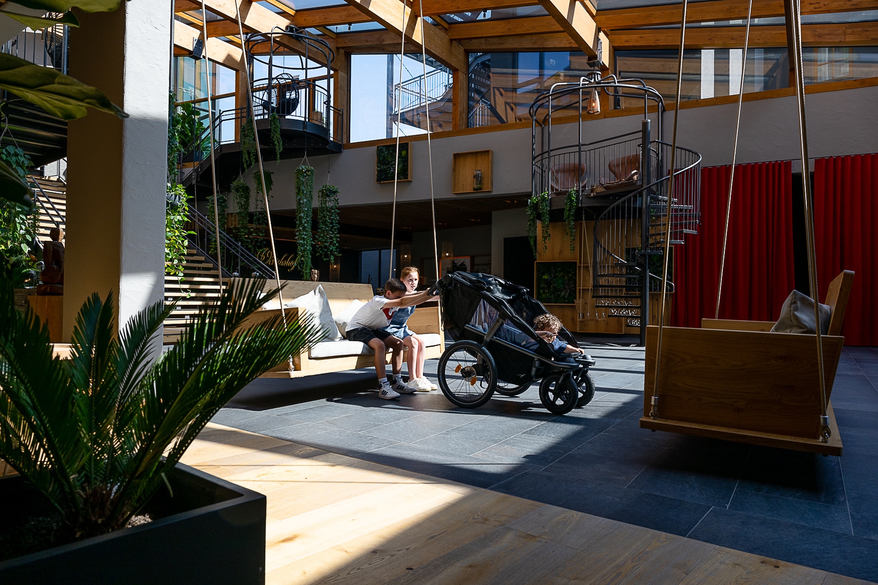 Zwei Kinder sitzen in einer Schaukel, vor ihnen steht ein Kinderwagen der Marke tfk im ULRICHSHOF Foyer